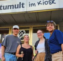 Vier Personen stehen vor einem Kiosk mit dem Banner "tumult im Kiosk" und lächeln in die Kamera.