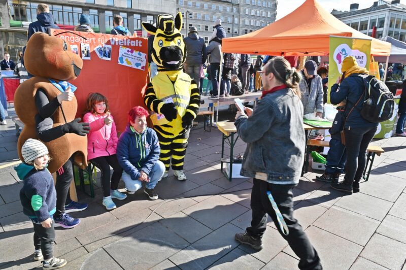 Drei Kinder stehen mit zwei Maskottchen vor einer Fotowand und lassen sich fotografieren.