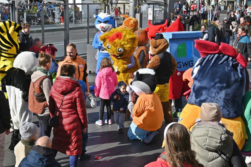 Eine Menschengruppe und einige Maskottchen stehen auf einem Marktplatz, unterhalten sich und machen Fotos zusammen.