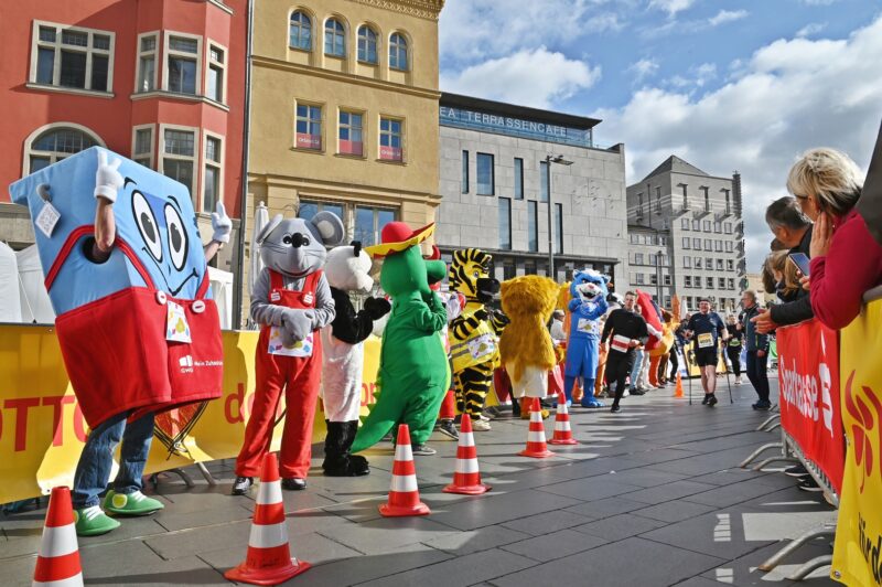 Eine Reihe von Maskottchen steht an einer Laufstecke und feuert laufende Personen an.