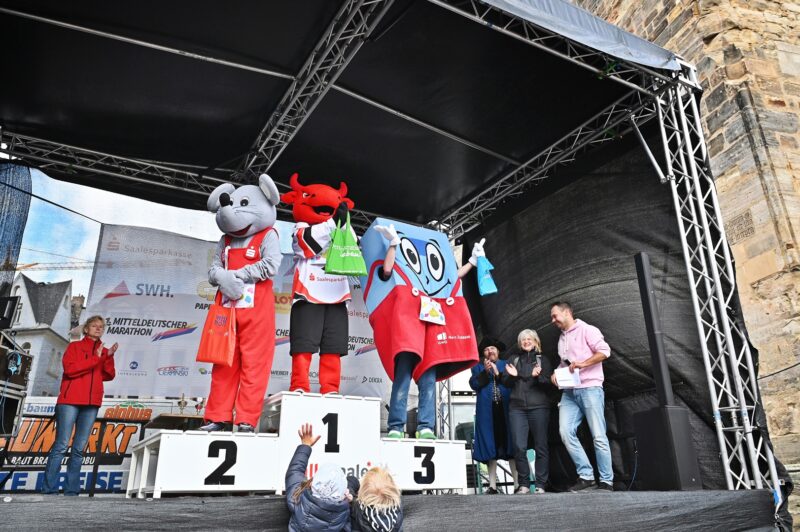 Drei Maskottchen stehen mit Medallien und Beuteln auf einer Sieger-Tribühne.