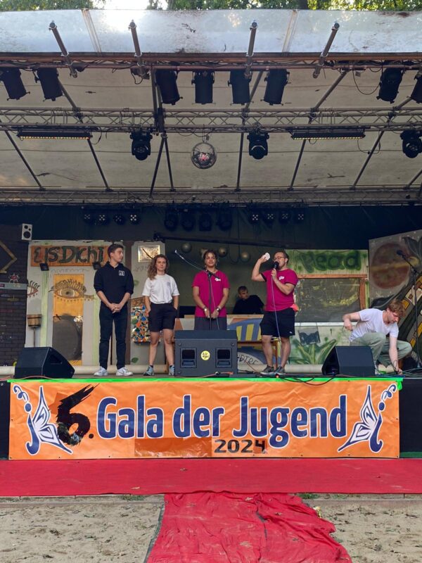 Vier Personen stehen auf einer Bühne mit dem Banner "6. Gala der Jugend". Die Person ganz rechts spricht in ein Mikrofon.