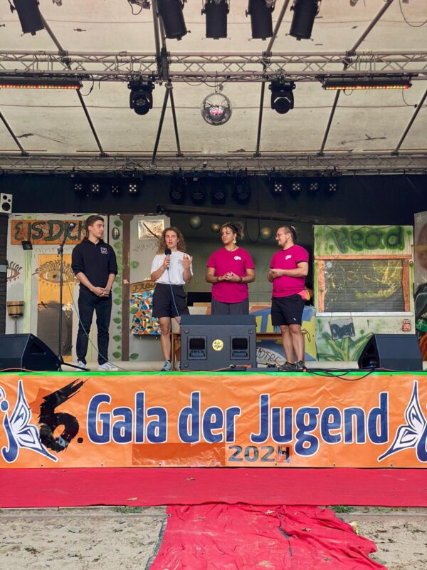 Vier Personen stehen auf einer Bühne mit dem Banner "6. Gala der Jugend". Eine von ihnen spricht in ein Mikrofon.